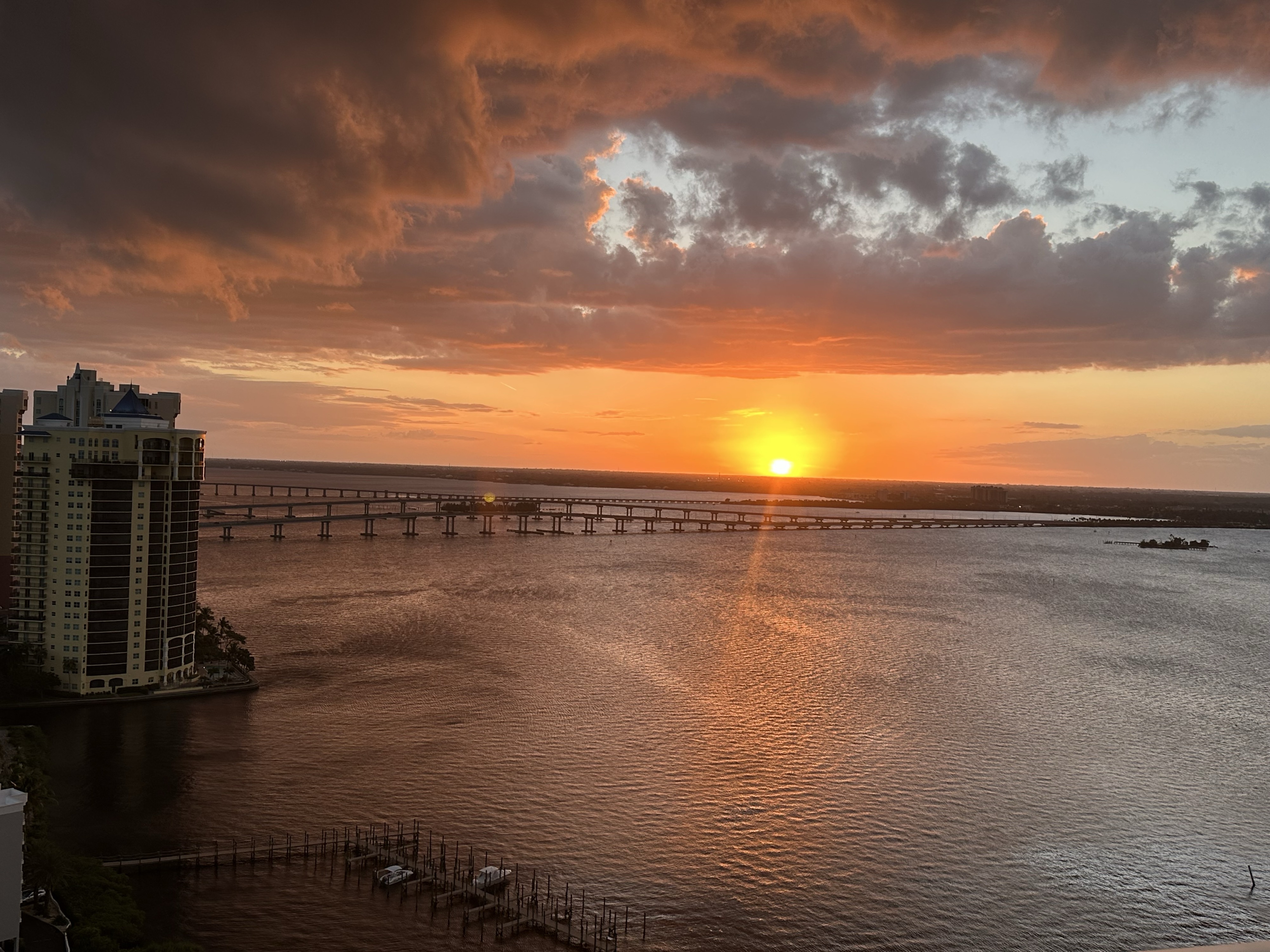 Orange sunset over dark river with big dramatic clouds. An orange beam or light shoots out of rh sun. The sun glows yellow with saturated heavy light around it.