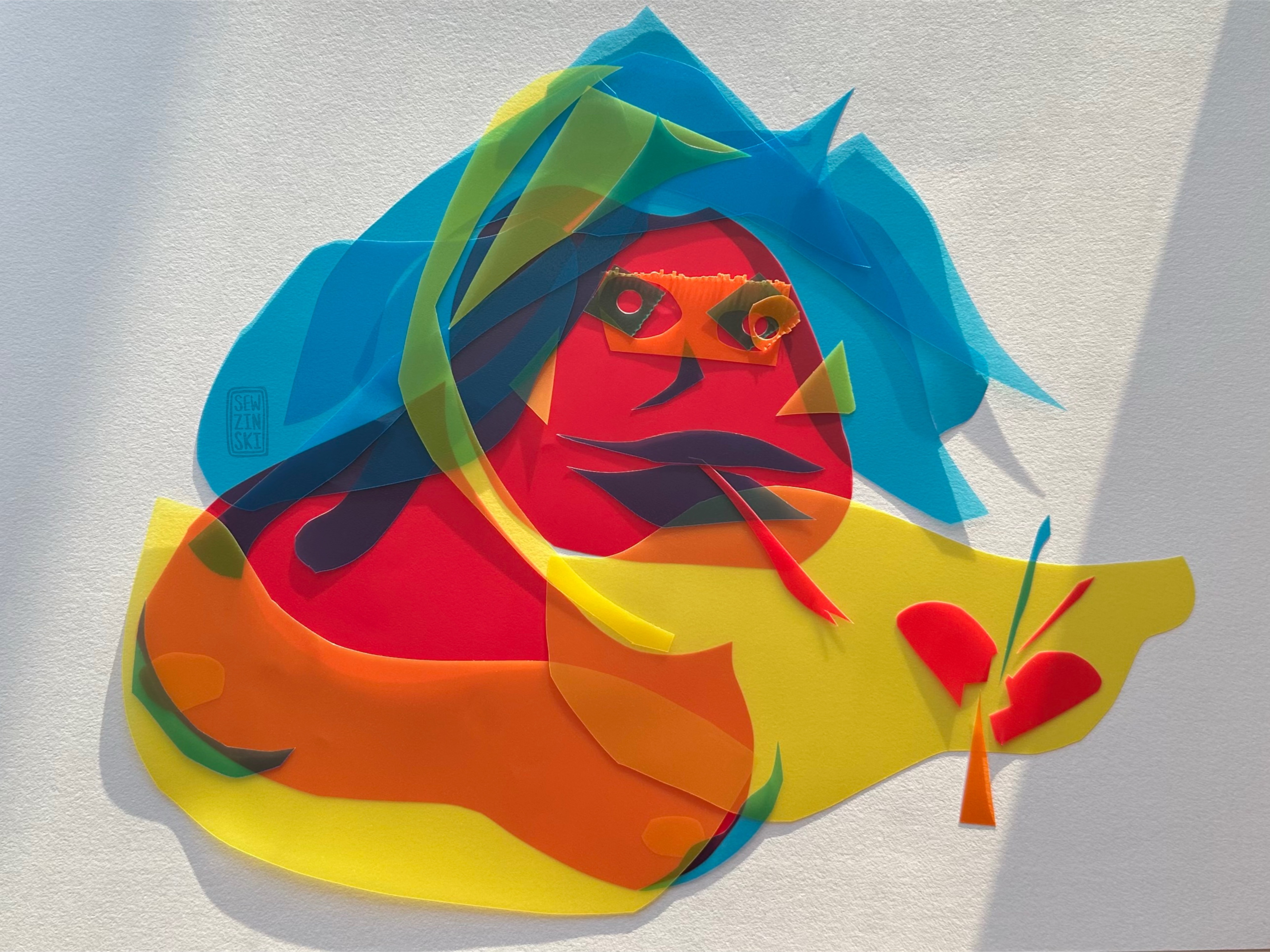 Vinyl I’ve shaped and layers into a woman looking creature with broken heart. She has a snake tongue, blue hair; and red skin with yellow arm wings.  Hand assembled collage with paper and plastic by me Sewzinski