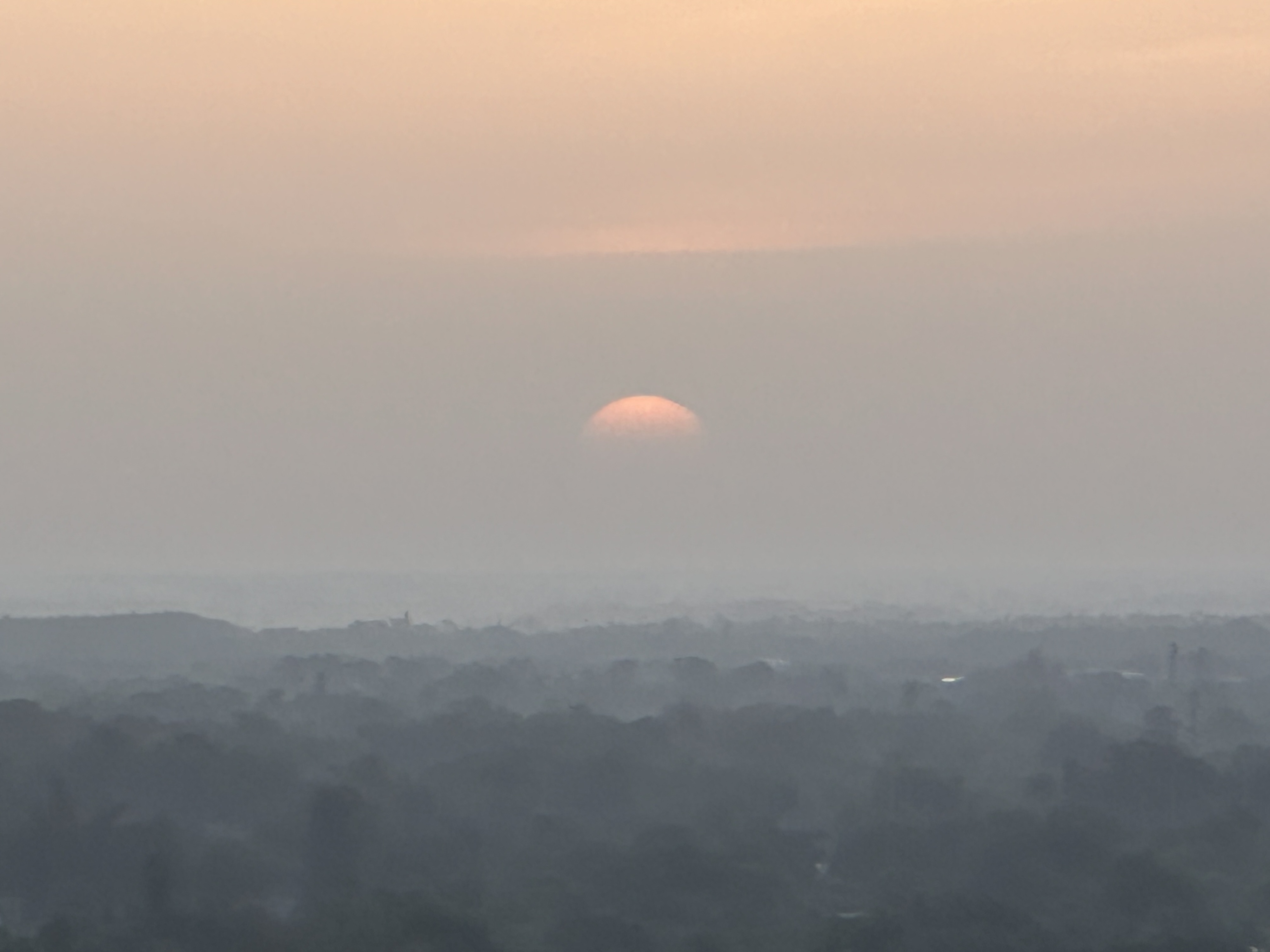 High pov: low flat landscape in fog and haze. Treetops can be vaguely seen through shadows. Off against a blue gray hazy background a small half ombre orange is rising.  iPhone no edits.