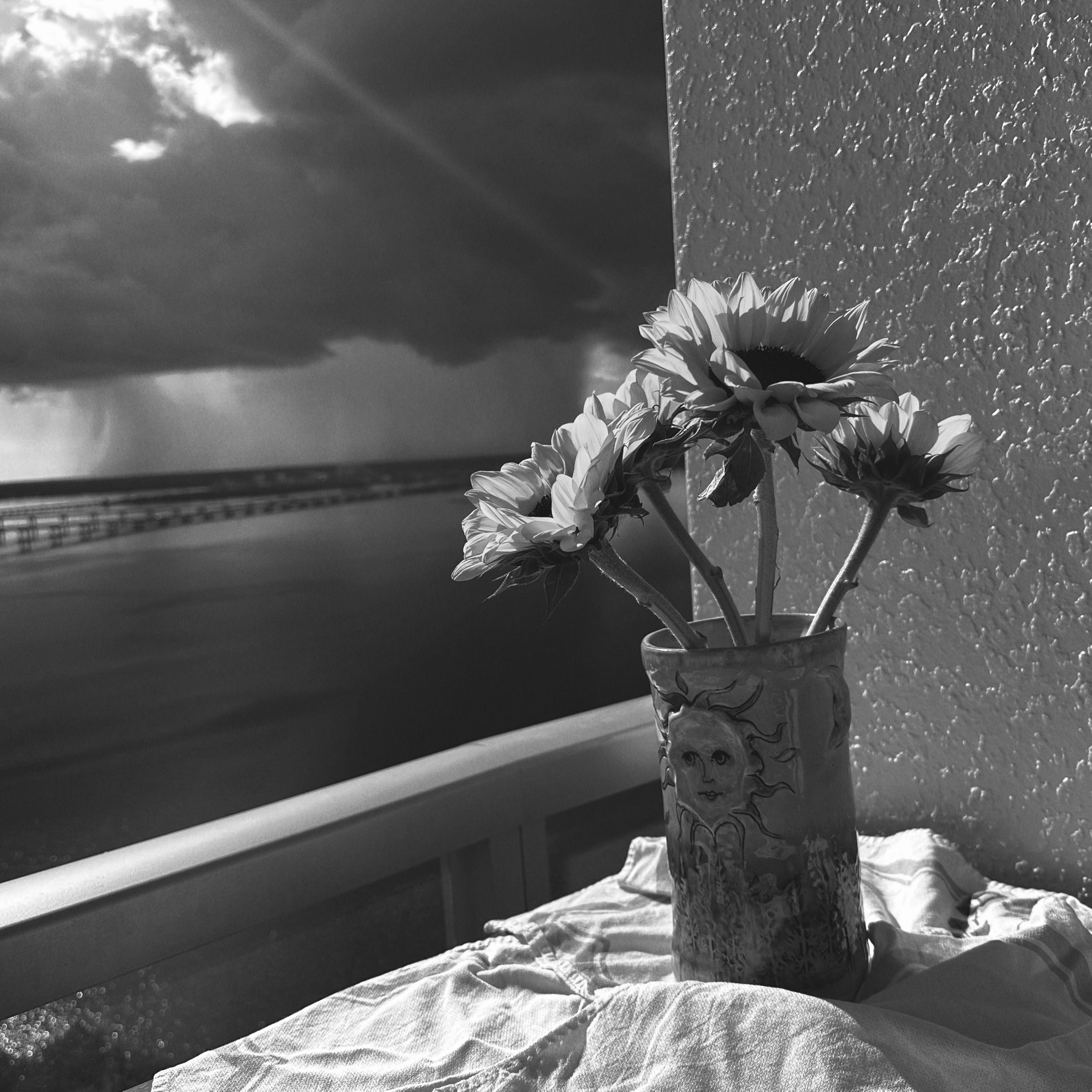 Sunflowers in a textured vase with a wide river and skinny bridges low and off in the distance. a stormy river and sky behind it. A beam of sunlight is falling towards the vase iPhone photo with filter.
