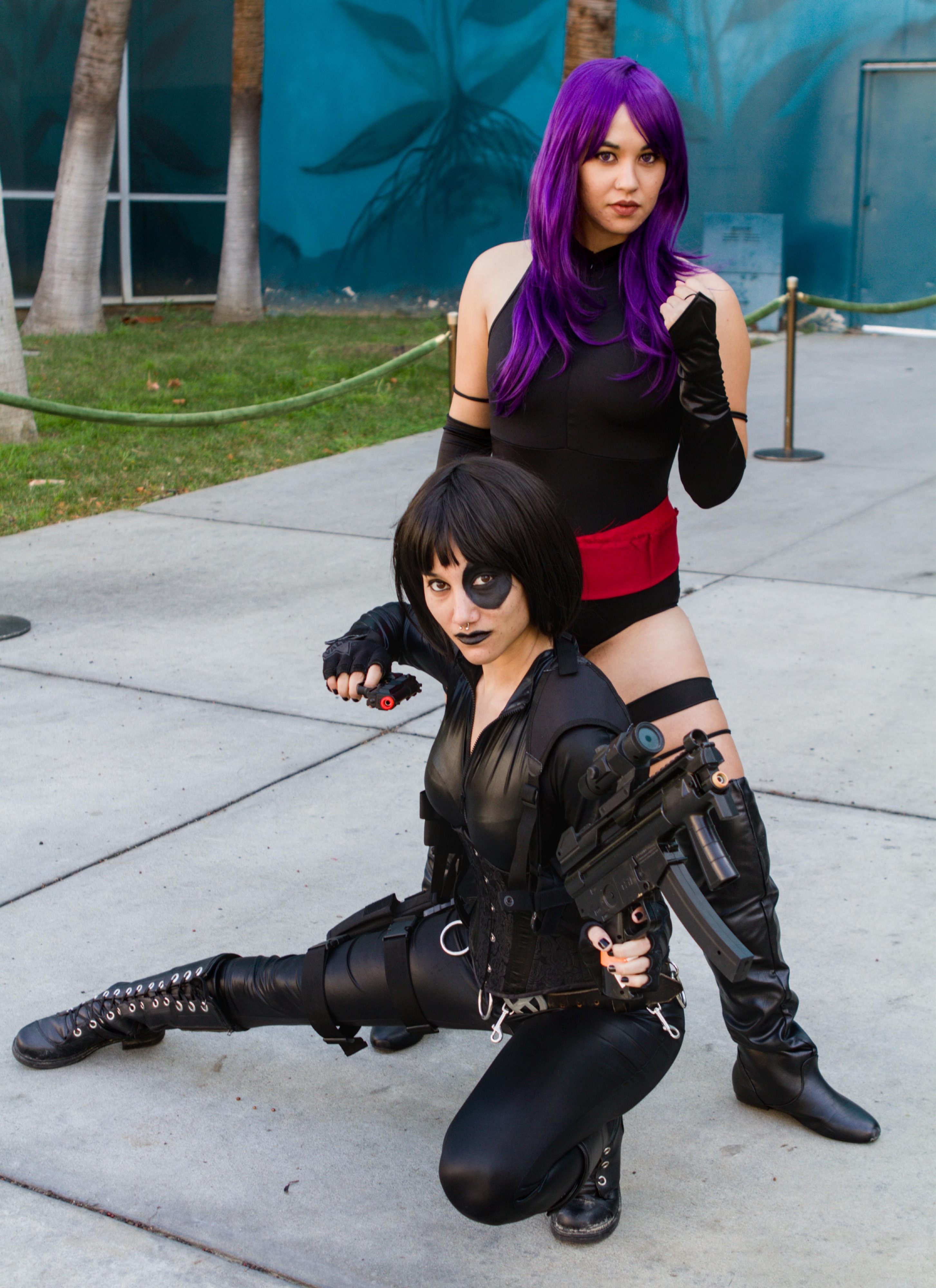 Two women cosplaying as the characters from the comic book X Men.  Psylocke is standing over Domino.  Psylocke has black boots, exposed thighs, black leotard with red sash at the hips and black leather bracers.  Her dark purple hair stands out as drapes across her breasts.

Domino has one leg kicked out and one leg bent below below her while aiming her guns at the camera.  She is covered all in black.  Her boots have a dozen or more metal lace up holes, which stand out along the front of the boot.  The outfit is skin tight and semi sheer at the breast.  Her hands are covered in fingerless gloves.
