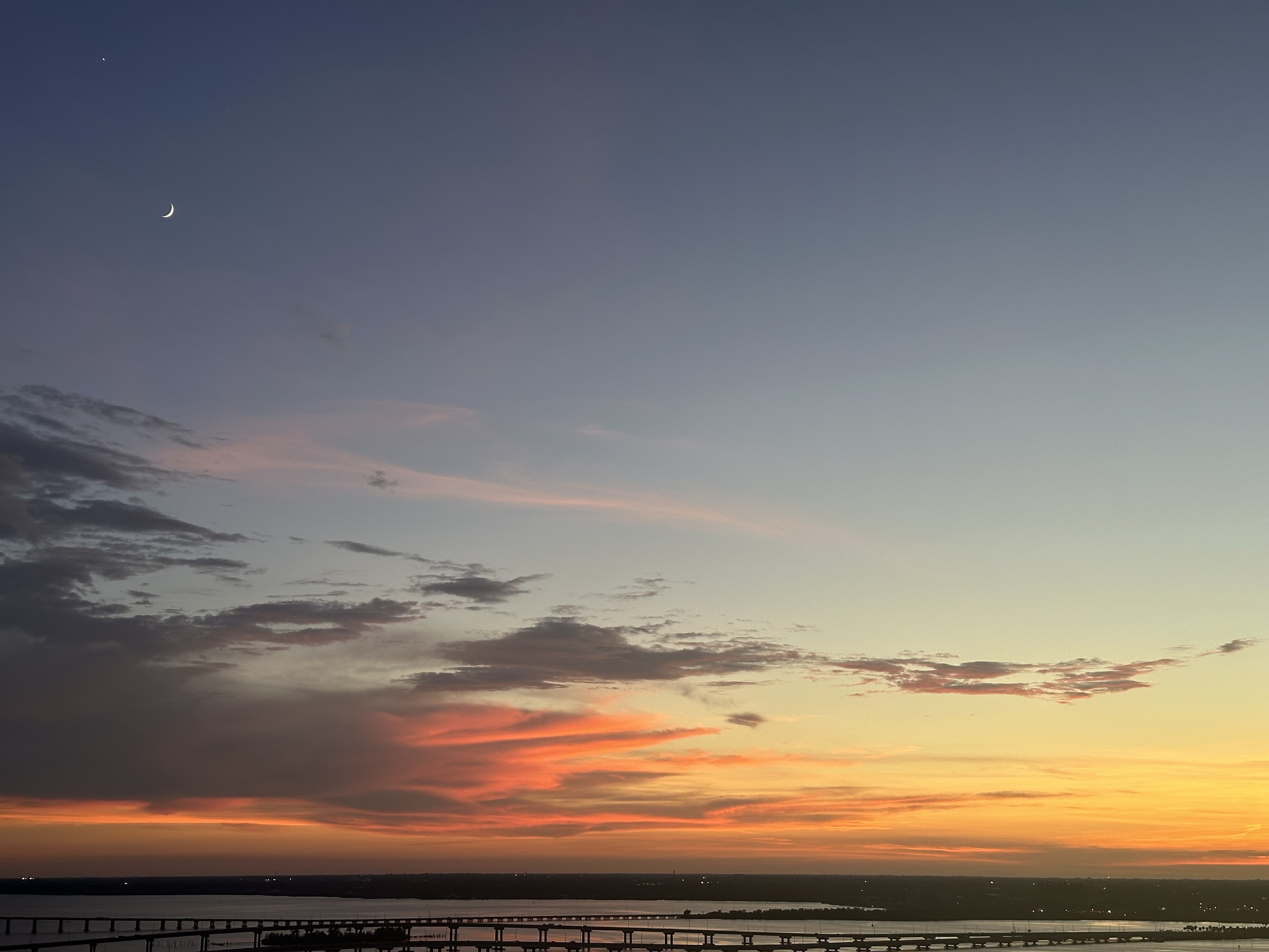 Warm orange and yellow clouds with gray and a blue dusk sky over river in shadow. The moon is a tiny crescent in the upper left with a star just above it bright. iPhone photograph