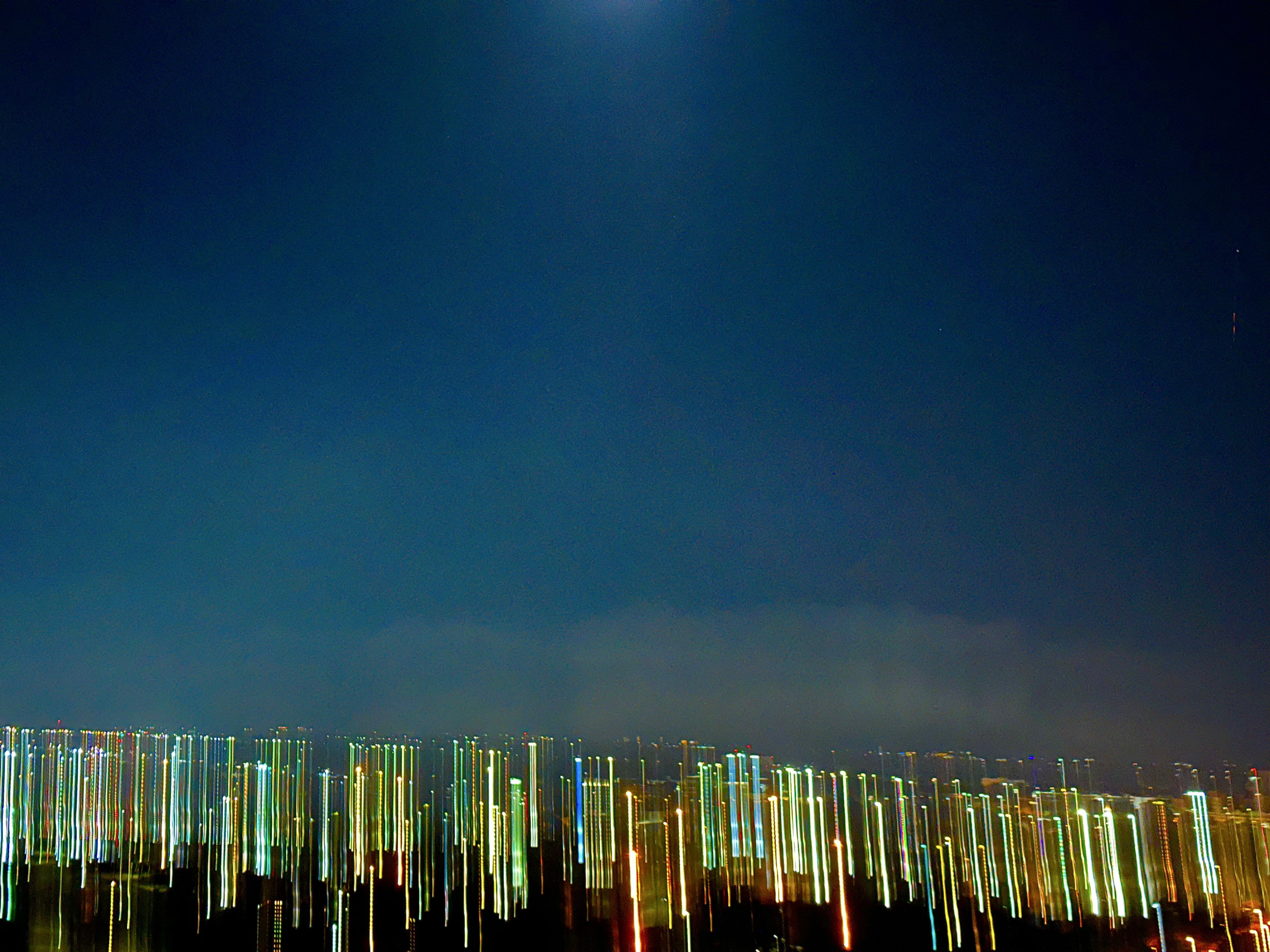 Deep teal sky has a beam of moonlight washing down towards city lights. No moon to be seen. City lights in blue, yellow, green, orange, smeared toward the ground which is in black shadow. It reminds me of the ocean with coral bottom.