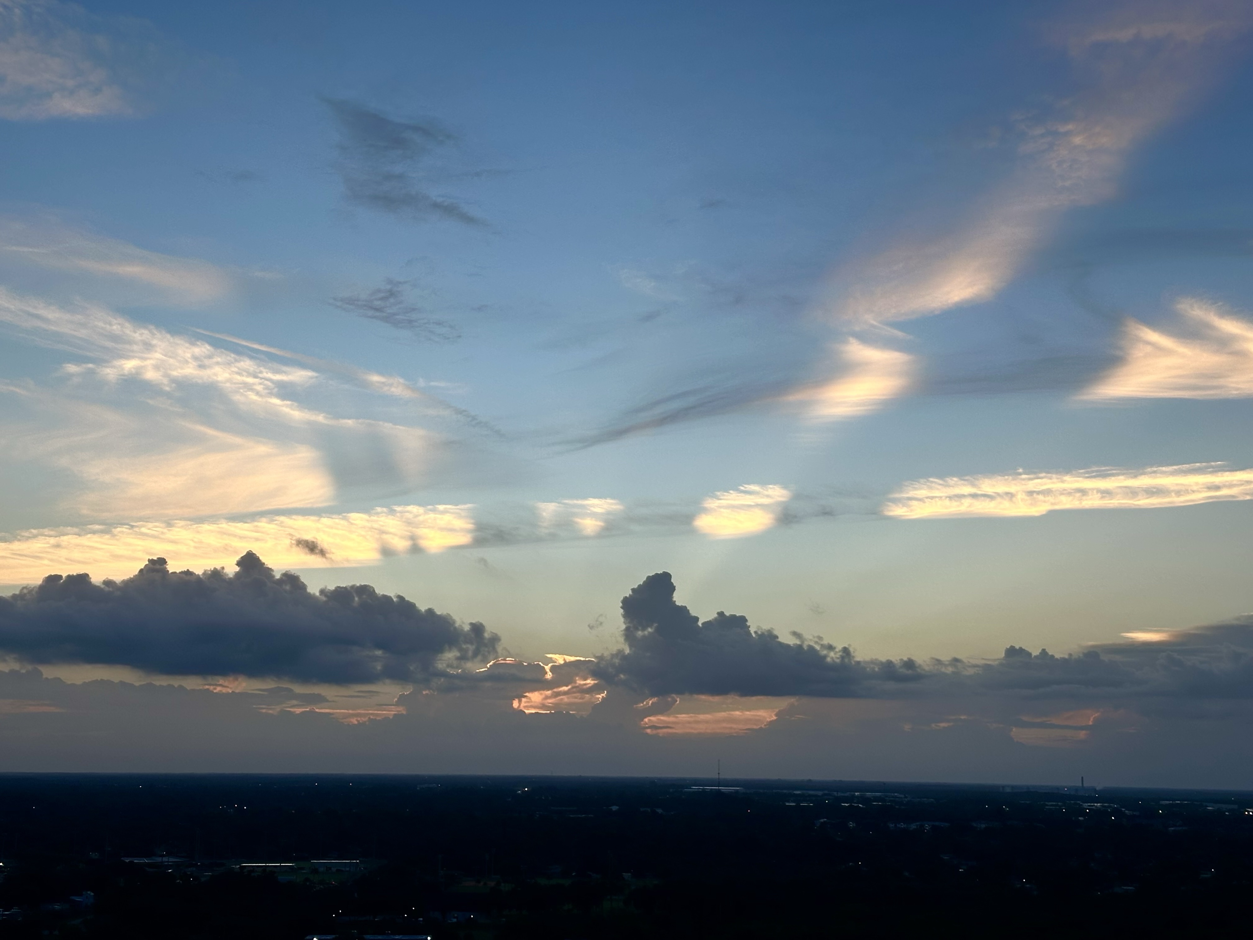 Low dark flat cityscape with a few visible lights. A blue cloud wall hugs the horizon. The sun is rising but can’t be seen yet. Light is cutting through giant clouds weaving shapes in the sky iPhone, minimal edits.