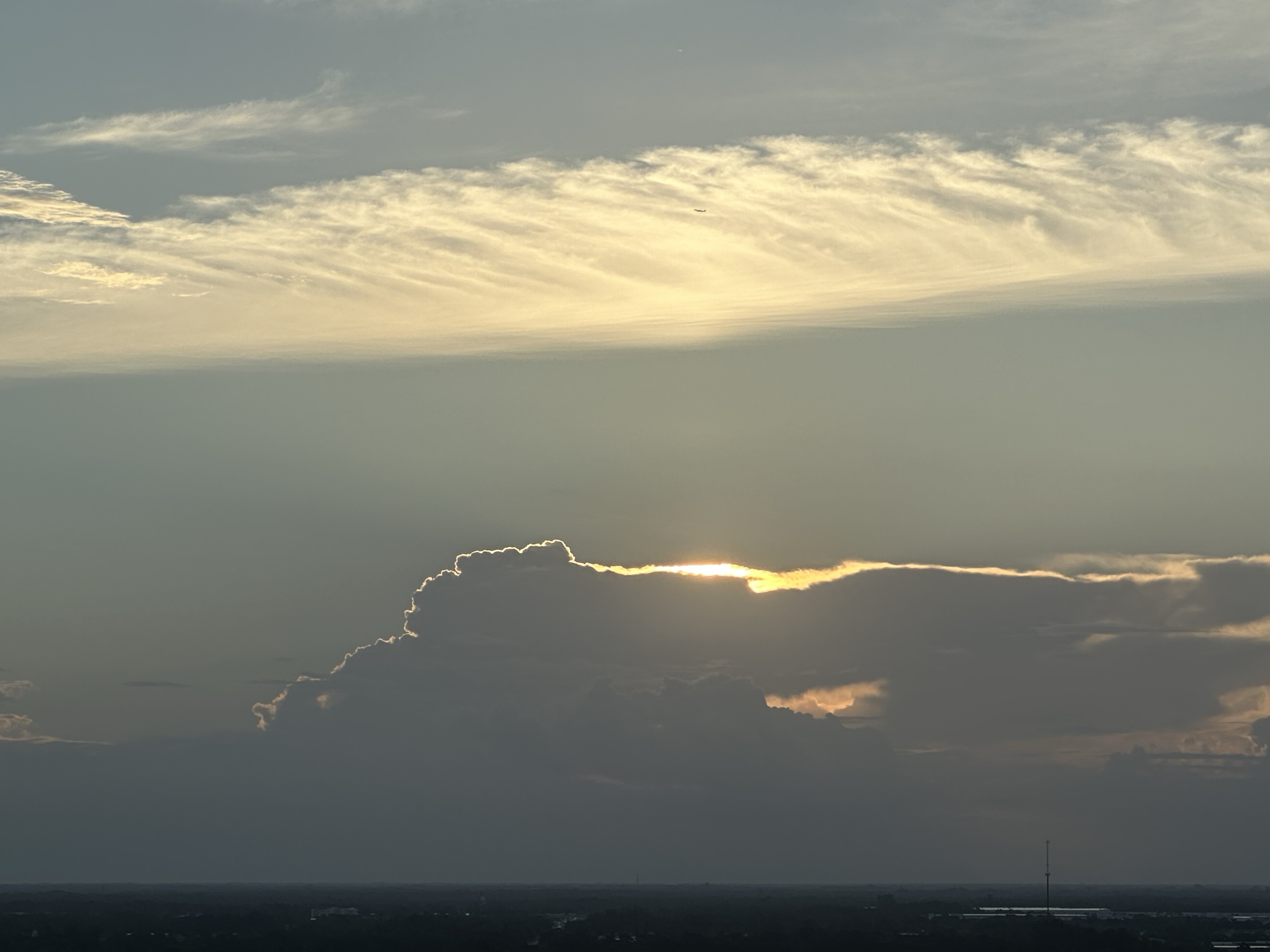 Long low flat lands in shadow. A wall of deep gray blue clouds covers the horizon where the sun is rising. It’s nearing the top of the clouds and a sliver of pure bright white light is hugging the curving sagging top of the clouds. The middle of the light line is white, but the edges fade to a yellow herein. The sky above is a hazy blue. A stripe of white clouds cuts horizontally like white crepe paper. iPhone, no edits.
